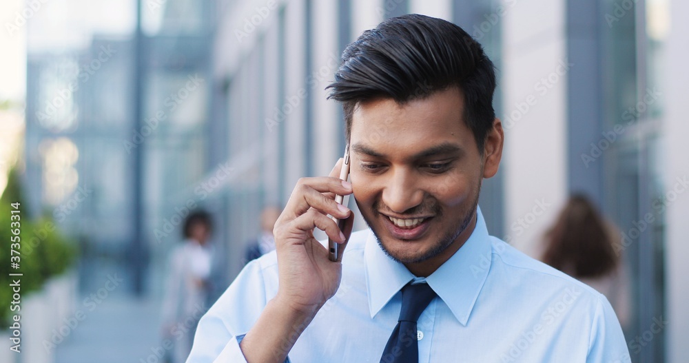 Hindu cheerful young businessman in tie standing at street and talking on phone. Man in business style speaking on mobile phone and laughing. Smiled happy male white-collar employee. Close up. Hindu cheerful young businessman in tie standing at street and talking on phone. Man in business style speaking on mobile phone and laughing. Smiled happy male white-collar employee. Close up.