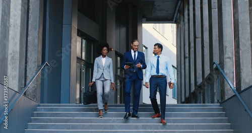 Mixed-races young men and woman leaving business office building and stepping down the steps. Outdoors. African American businesswoman and Caucasian Hindu businessmen talking and using tablet device.