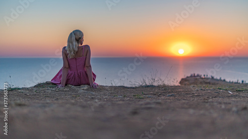 Smiling young woman with charming smile looks at the sunset stands with her back in the summer in the evening