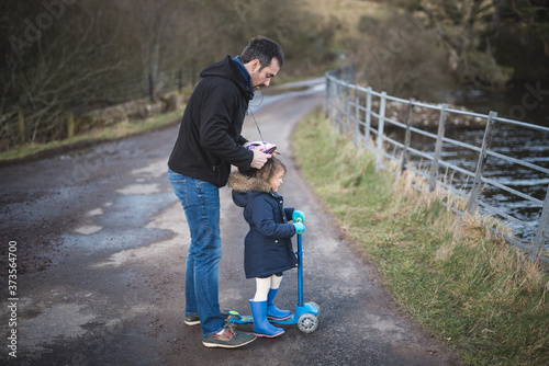 A father helps her daughter by putting on her helmet before riding her scooter on a road beside a lake before sunset in the Pentland hills regional park in Edinburgh, Scotland, United Kingdom photo