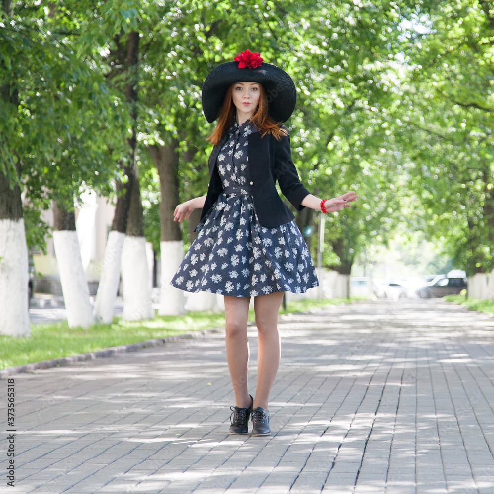 Redhead girl in vintage dress and wide-brimmed hat dances on alley in summer park