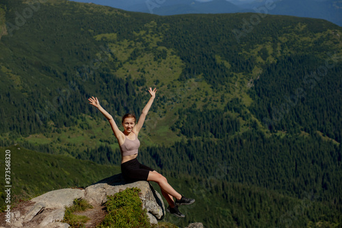 The young girl sitting at the top of the mountain raised her hands up on forest background. The woman climbed to the top and enjoyed her success.
