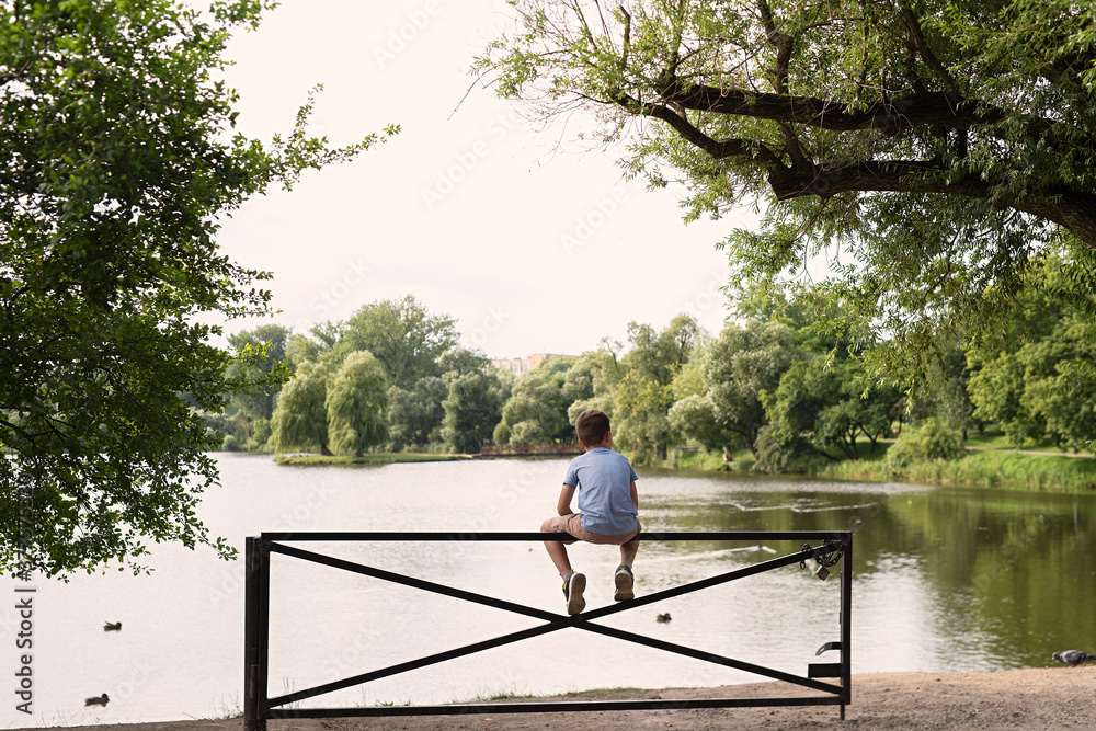 boy sits on  fence and admires  picturesque nature, trees and river on  background