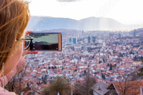 Girl with glasess and brown hair photographing cityscape of Sarajevo capital of BiH with mobile phone photo