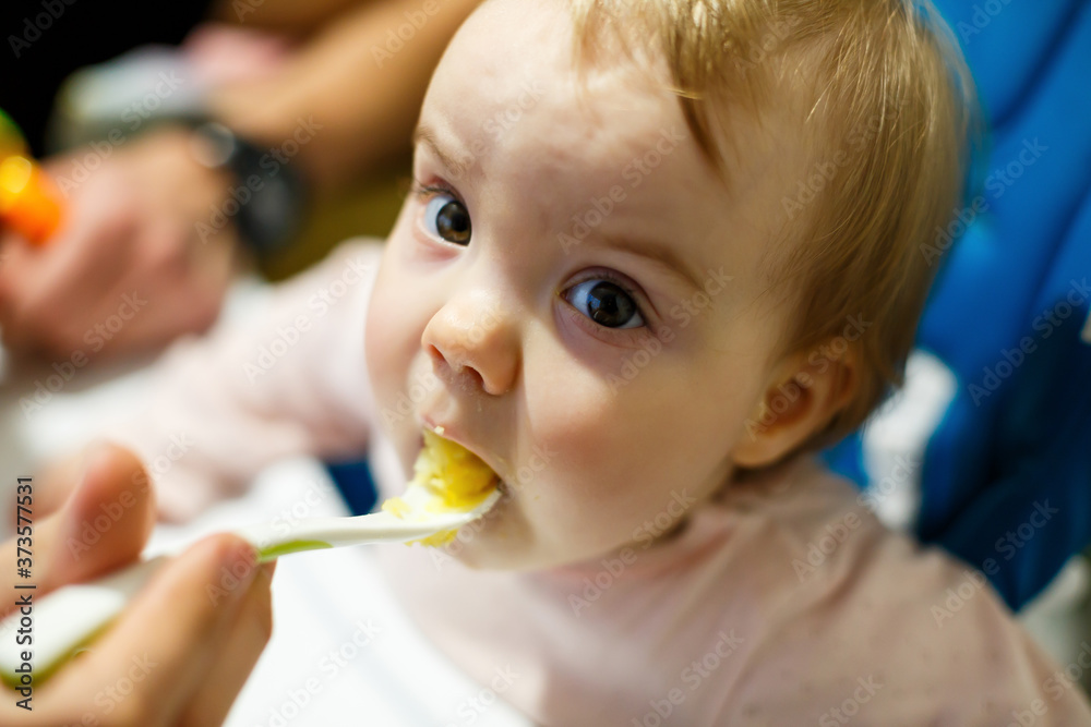 Photo portrait of a baby girl with pink cheeks she sits in a chair for feeding and eats porridge. Child sitting at home Photo portrait of a baby girl with pink cheeks she sits in a chair for feeding and eats porridge. Child sitting at home