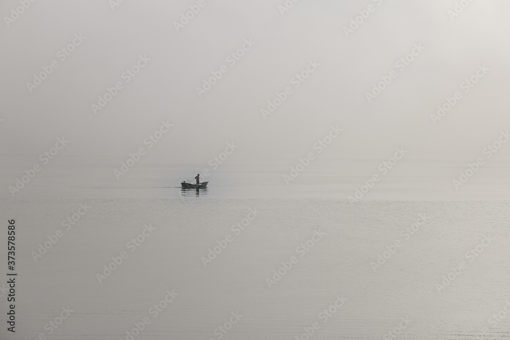 Fototapeta premium Homem navegando num pequeno barco com mar e céu encobertos pela névoa
