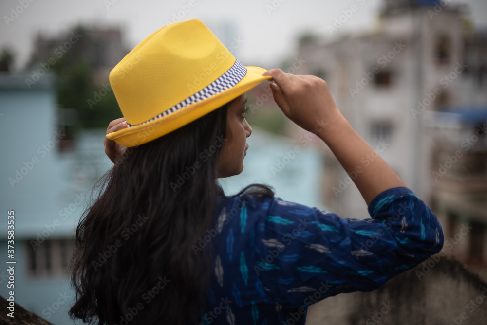 Backside view of an Indian woman standing on rooftop wearing a blue dress and yellow western hat in a cloudy afternoon. Indian lifestyle Backside view of an Indian woman standing on rooftop wearing a blue dress and yellow western hat in a cloudy afternoon. Indian lifestyle