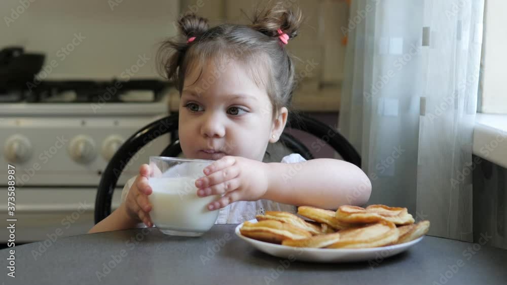 Adorable little girl is eating cheesecakes with her hands at the table in kitchen. Glass of yogurt on the table Adorable little girl is eating cheesecakes with her hands at the table in kitchen. Glass of yogurt on the table