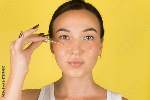 A beautiful young girl applies a serum to her face with a pipette. Cosmetology, skin care. Yellow background