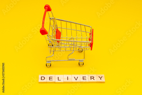 Word DELIVERY made from wooden cubes on yellow background with toys shopping cart
