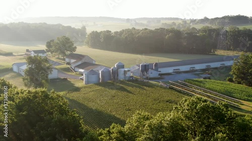 Establishing shot, aerial of family farm in United States, grain bins, chicken house, barn and fields in summer hazy foggy morning light