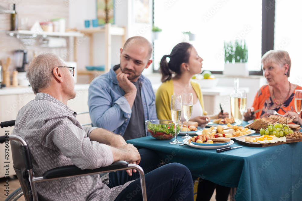Disabled senior in wheelchair man having a conversation with son during family brunch in kitchen. Senior parents together with mature children.