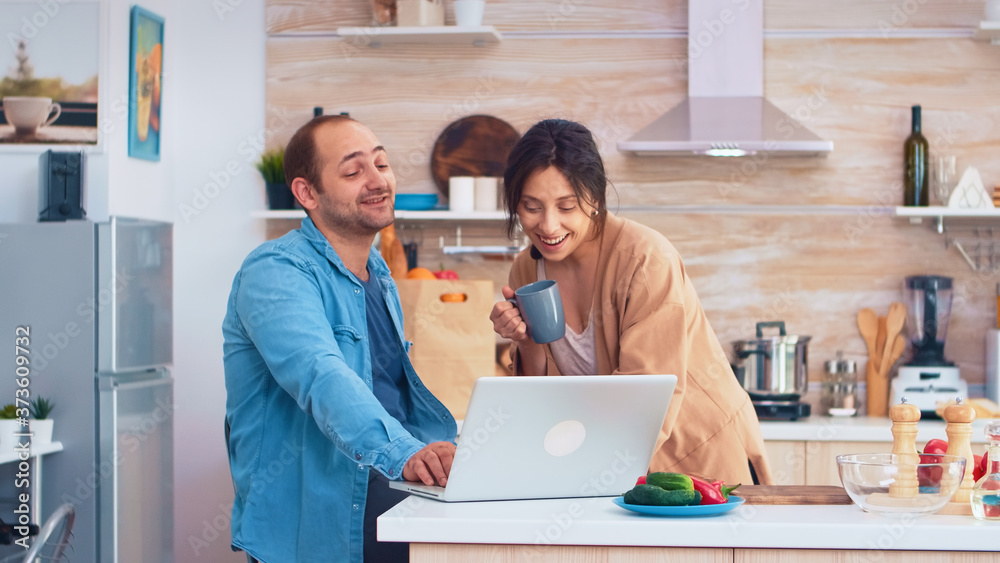 Entrepreneur working on laptop in kitchen and wife holding a cup of coffee looking at his work. Husband and wife cooking recipe food. Happy healthy together lifestyle. Family searching for online meal Entrepreneur working on laptop in kitchen and wife holding a cup of coffee looking at his work. Husband and wife cooking recipe food. Happy healthy together lifestyle. Family searching for online meal