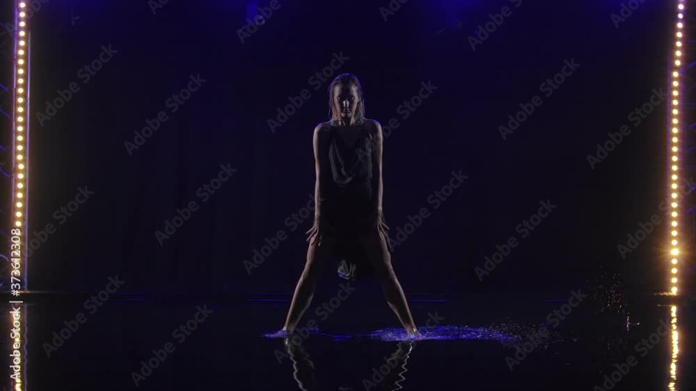 Silhouette of a woman performing an element of bachata dance under rain. Blonde dances on the surface water creating lots of splashes. Black background with neon spotlights. Slow motion. Silhouette of a woman performing an element of bachata dance under rain. Blonde dances on the surface water creating lots of splashes. Black background with neon spotlights. Slow motion.