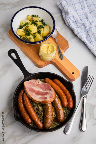 Oldenburger Grünkohl mit Pinkel, Kohlwurst, Kassler Schweine Nacken, Senf und Kartoffeln in Gusseisen Pfanne auf Marmor Tisch