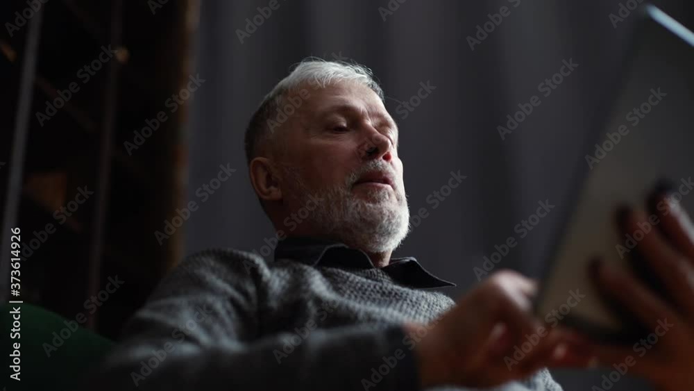 Close-up view from below of handsome mature adult male having conversation by digital tablet. Older man alone at home has video call on tablet to keep in touch with family during isolation. Close-up view from below of handsome mature adult male having conversation by digital tablet. Older man alone at home has video call on tablet to keep in touch with family during isolation.