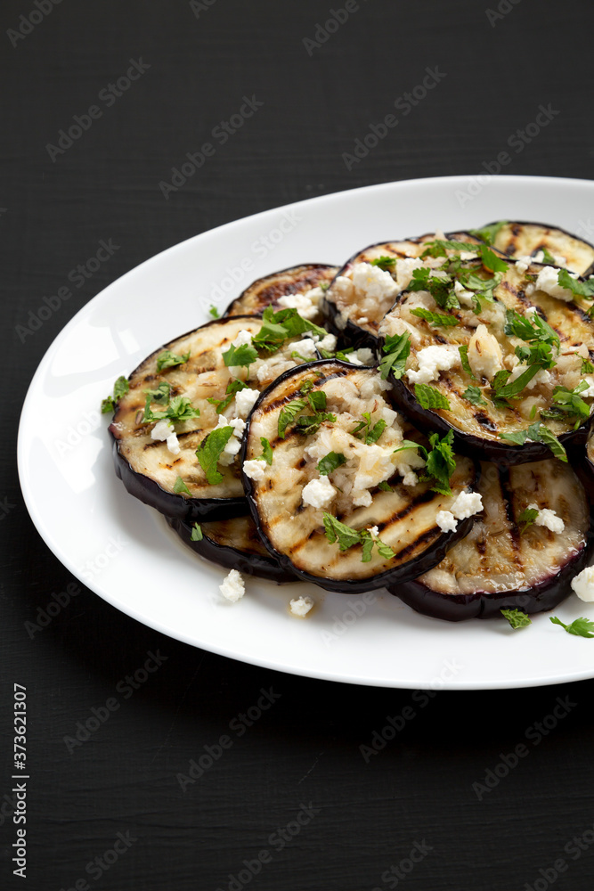 Homemade Grilled Eggplant with Feta and Herbs on a white plate on a black background, side view. Homemade Grilled Eggplant with Feta and Herbs on a white plate on a black background, side view.