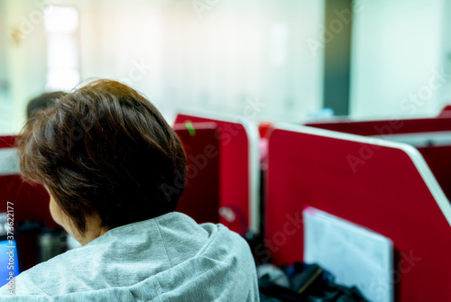 Back view of short hair Asian woman in office. Woman worker sitting at her desk in office. People work at her red table at workplace. Women worker in casual lifestyle working with routine work.