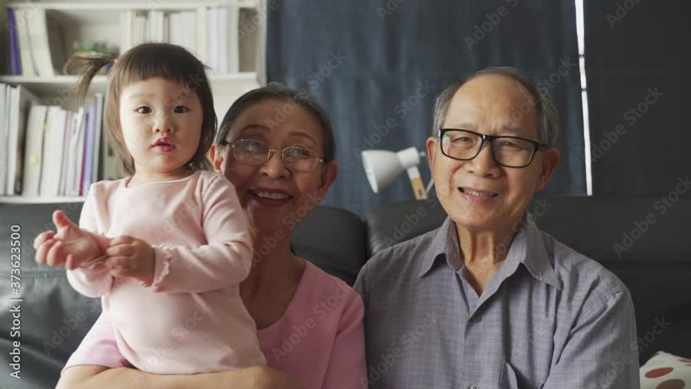 Asian family in living room, grandparents and little young nephew girl playing and smiling to camera. Happiness and enjoy lifestyle in home. Grandmother hug lovely cute kid baby sitting on sofa.