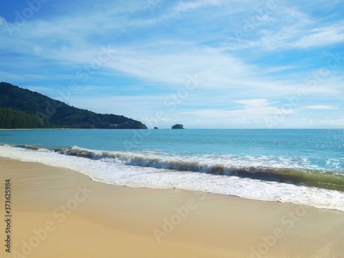 Coastal waves wash a white sand. Sea foam on the crest of the wave. Empty beach. Panoramic views of the open ocean and the green promontory stretching to a horizon. Thailand. Phuket. Tropical paradise
