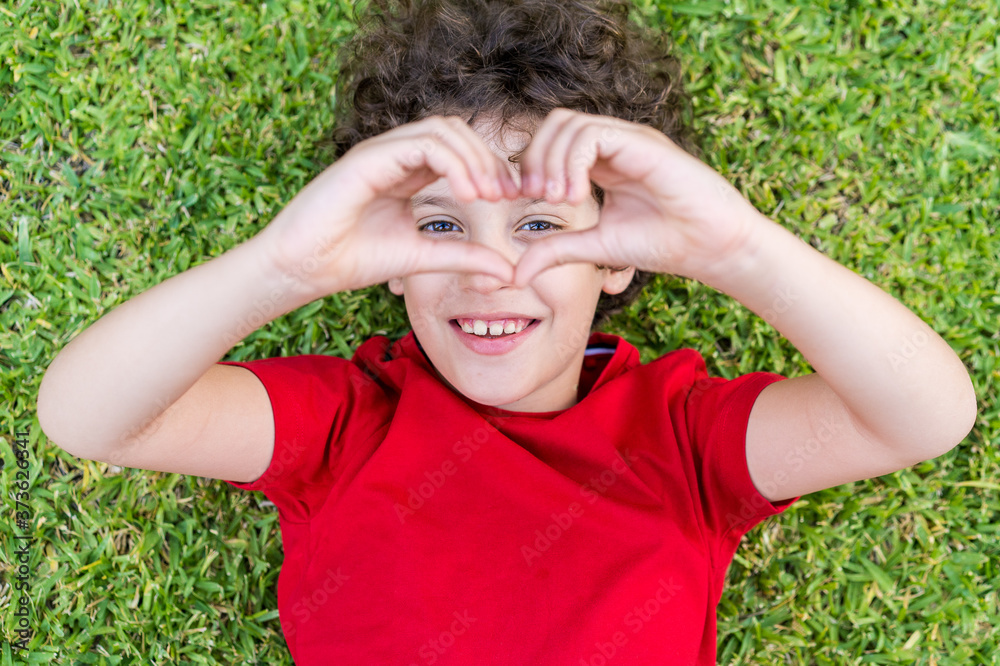 Cute kid lying in the grass and making a love heart symbol with the fingers. Top down lifestyle portrait. Cute kid lying in the grass and making a love heart symbol with the fingers. Top down lifestyle portrait.