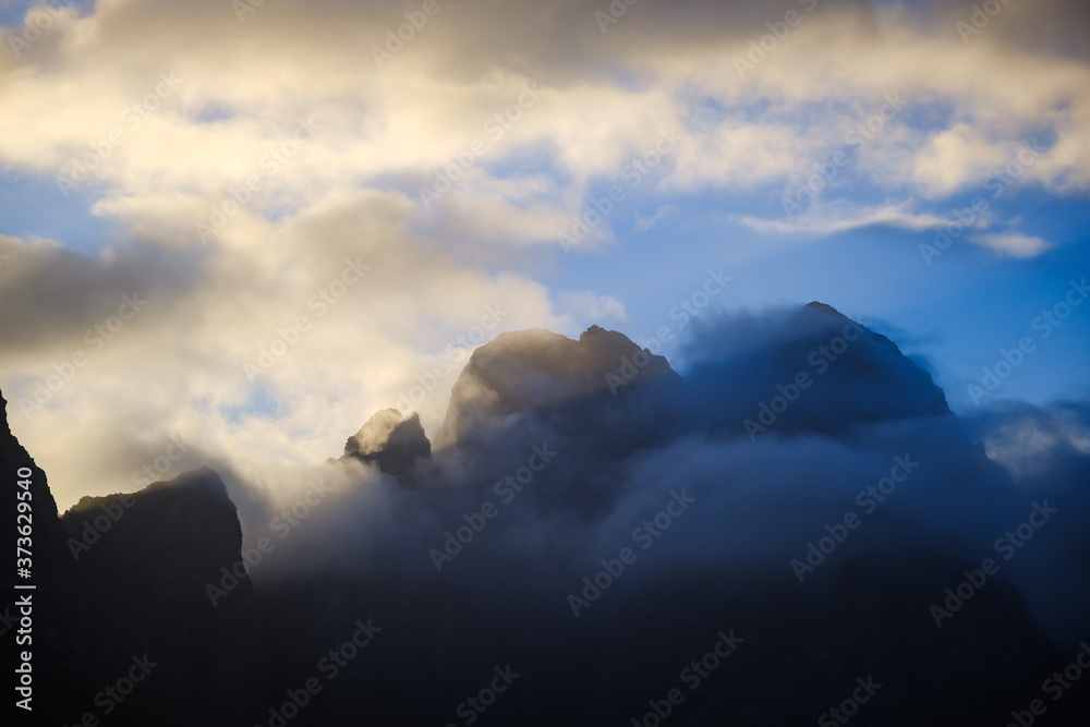 Sun rising behind jagged mountain summit and swirling clouds landscape.