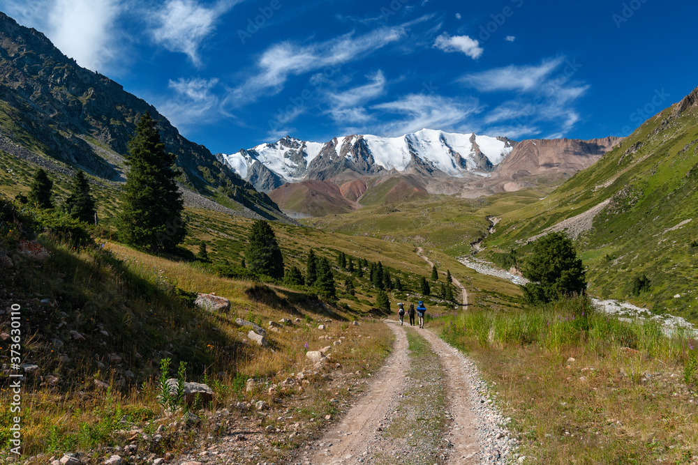 Scenic landscape with a group of three unidentified tourists with backpacks walking along the road towards the snowy mountains, Tien Shan, Kazakhstan