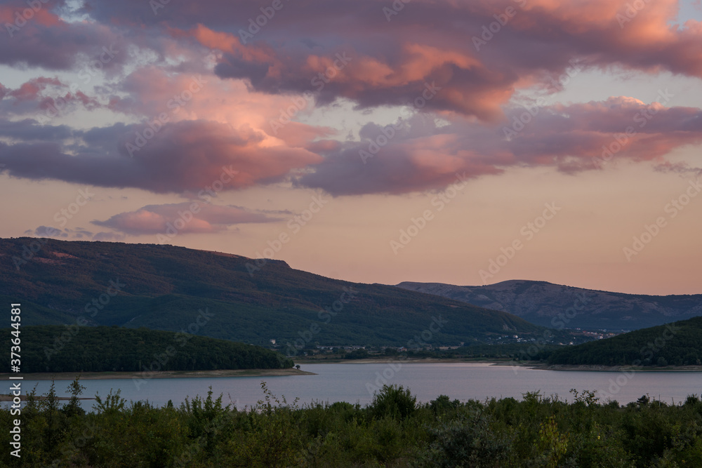 Pink clouds over the lake and forest. Bright sunset light. Purple and crimson shades. Rural landscape at sunset. A rustic motif with a lake and mountains.