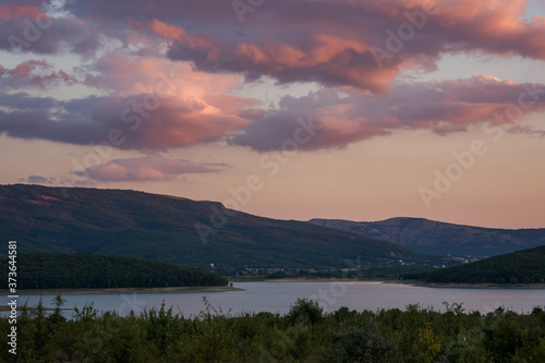 Pink clouds over the lake and forest. Bright sunset light. Purple and crimson shades. Rural landscape at sunset. A rustic motif with a lake and mountains.