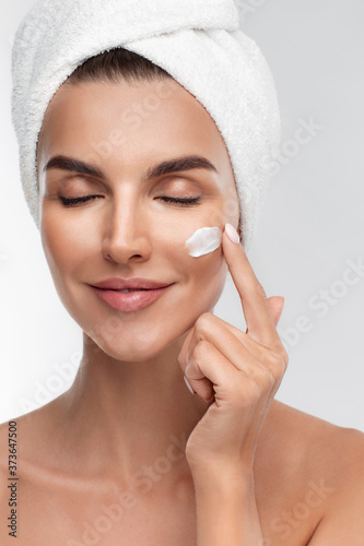 Close up portrait of half naked young woman applying moisturizing face anti-aging cream on flawless skin, closed eyes, isolated on grey background. Natural makeup, touching face. Beauty routine
