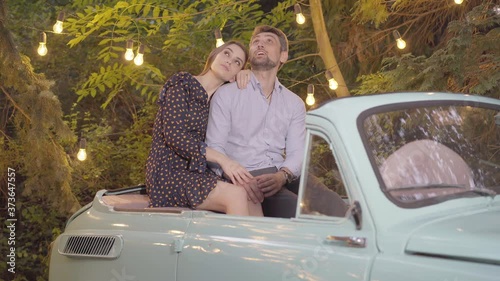 Camera approaches to relaxed loving couple sitting in retro car and chatting. Portrrait of happy Caucasian man and woman dating in summer evening outdoors. Romance concept. photo