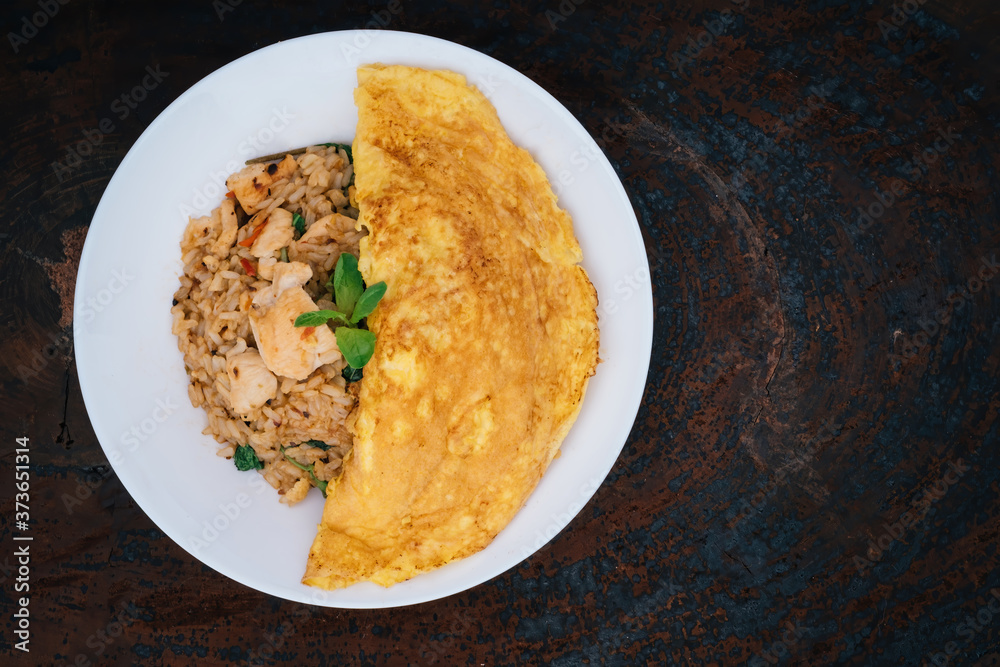 homemade Thai style spicy fried rice with stir-fried chicken and basil cover by a half circle omelet in white plate with spoon in white napkin on wood Serving trays with dark gray background. homemade Thai style spicy fried rice with stir-fried chicken and basil cover by a half circle omelet in white plate with spoon in white napkin on wood Serving trays with dark gray background.