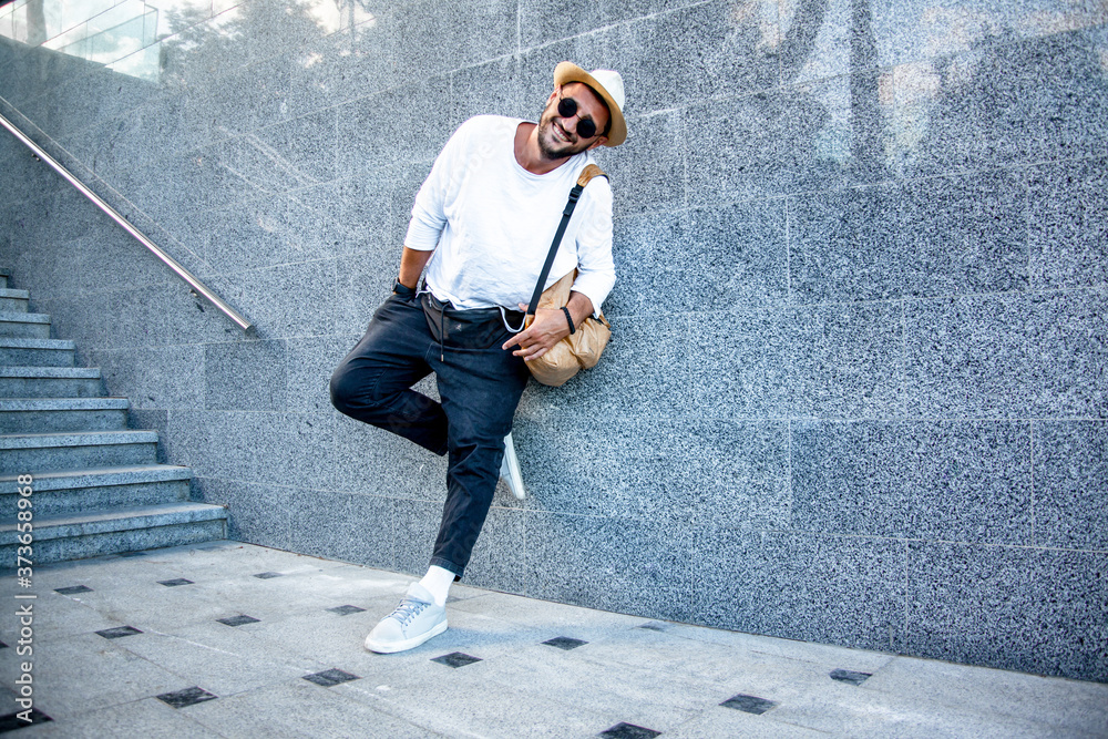 Portrait of stylish handsome man with a beard dressed white t-shirt, black sunglasses, jeans with bagpack posing against the urban wall in the city on the street Portrait of stylish handsome man with a beard dressed white t-shirt, black sunglasses, jeans with bagpack posing against the urban wall in the city on the street