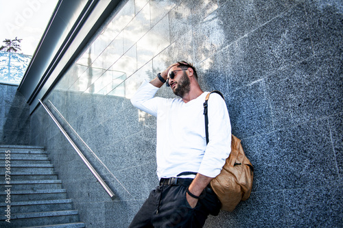 Portrait of stylish handsome man with a beard dressed white t-shirt, black sunglasses, jeans with bagpack posing against the urban wall in the city on the street