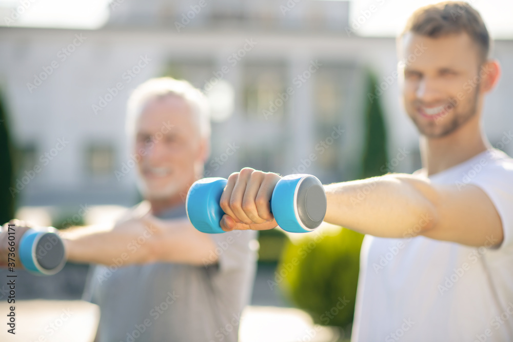 Young bearded male and grey-haired man practicing with blue dumbbells outside, smiling Young bearded male and grey-haired man practicing with blue dumbbells outside, smiling