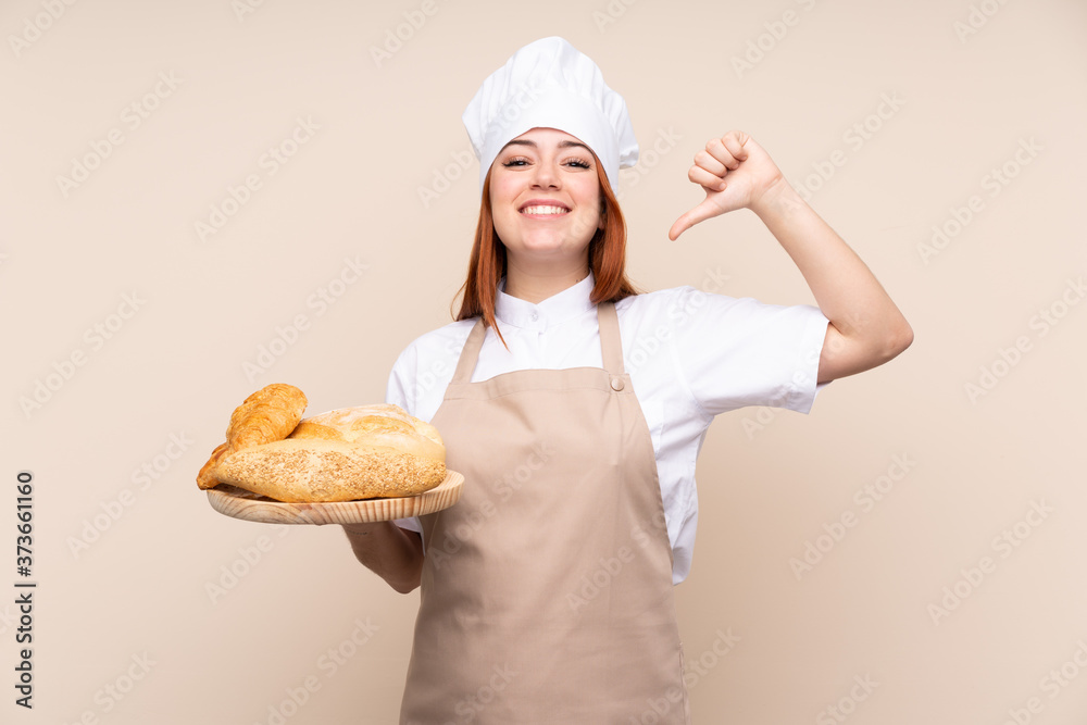 Redhead teenager girl in chef uniform. Female baker holding a table with several breads proud and self-satisfied