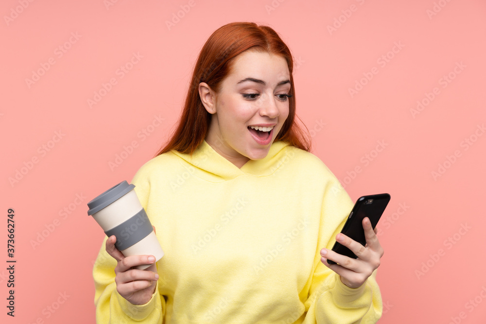 Redhead teenager girl over isolated pink background holding coffee to take away and a mobile Redhead teenager girl over isolated pink background holding coffee to take away and a mobile
