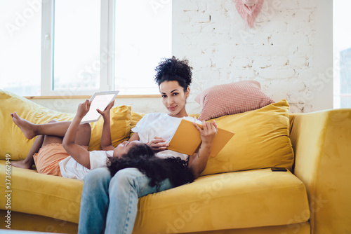 Relaxing woman and girl on bright couch with book and tablet