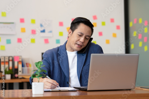 Business Asian man talking job on smart phone and note job in office