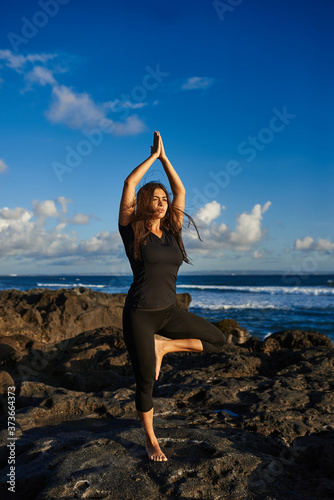 The girl practices yoga on the rocks overlooking the sea and blue sky..