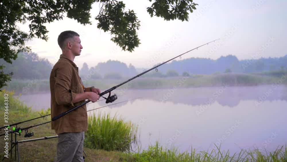 Silhouette a man. young male fisherman stands with a spinning fishing rod by the lake. The guy catches a fish holding a reel. night or morning fog over water, fish tackle. Camping and favorite hobby Silhouette a man. young male fisherman stands with a spinning fishing rod by the lake. The guy catches a fish holding a reel. night or morning fog over water, fish tackle. Camping and favorite hobby