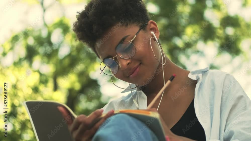 A smiling young african american woman with earphones is writing in her notebook sitting in the park