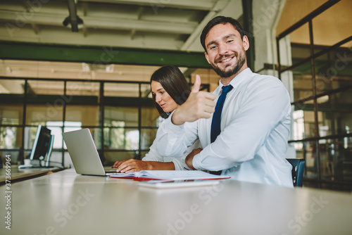 Portrait of successful male professional in formal wear enjoying work day in office, smiling Caucasian male employee looking at camera and showing ok while female colleague searching web information