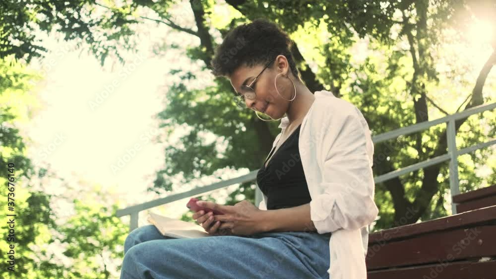A focused young african american woman with eyeglasses is using her smartphone sitting on the bench in the park A focused young african american woman with eyeglasses is using her smartphone sitting on the bench in the park