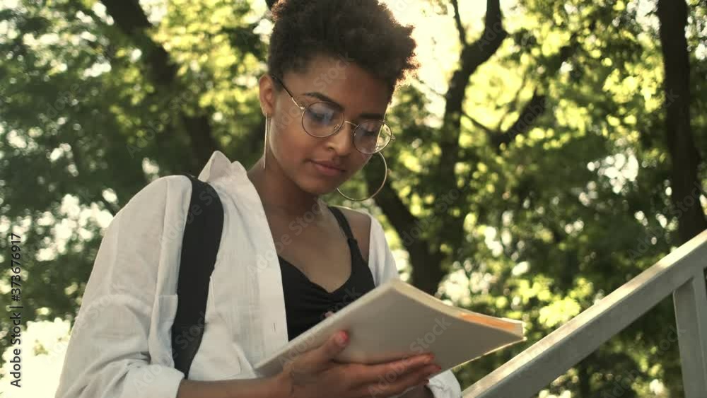 A nice focused young african american woman is looking in her notebook walking in the park near the bench A nice focused young african american woman is looking in her notebook walking in the park near the bench