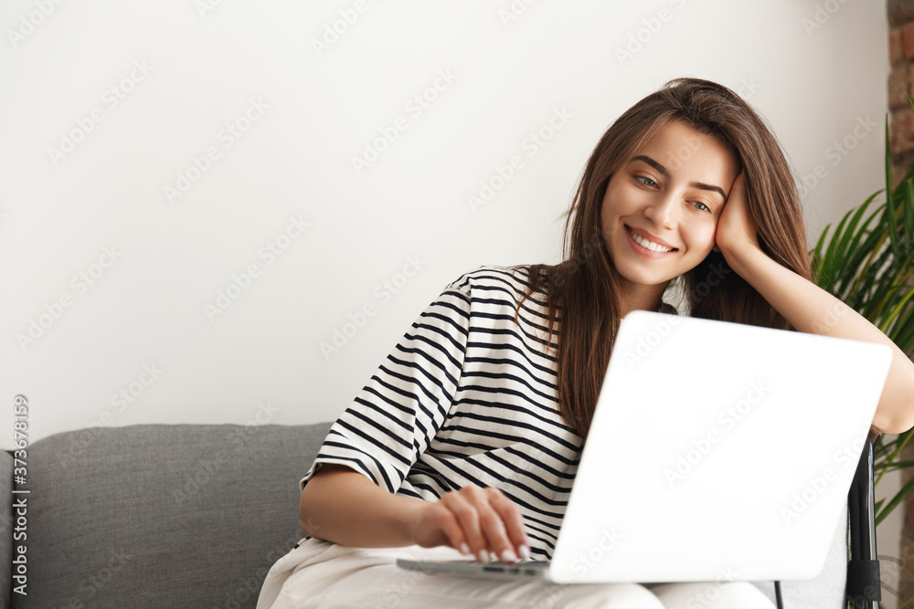 Smiling attractive girl relaxing on sofa, looking at laptop screen. Woman lying on couch and watching movies on computer. Student browsing social network, shopping online Smiling attractive girl relaxing on sofa, looking at laptop screen. Woman lying on couch and watching movies on computer. Student browsing social network, shopping online