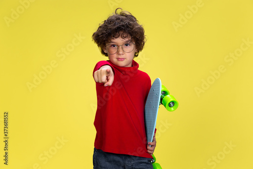 Pointing. Portrait of pretty young curly boy in red wear on yellow studio background. Childhood, expression, education, fun concept. Preschooler with bright facial expression and sincere emotions.