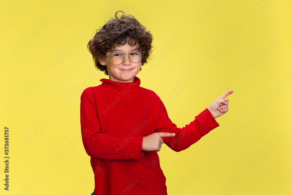 Pointing. Portrait of pretty young curly boy in red wear on yellow studio background. Childhood, expression, education, fun concept. Preschooler with bright facial expression and sincere emotions. Pointing. Portrait of pretty young curly boy in red wear on yellow studio background. Childhood, expression, education, fun concept. Preschooler with bright facial expression and sincere emotions.