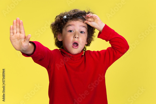 Shocked stopping. Portrait of pretty young curly boy in red wear on yellow studio background. Childhood, expression, education, fun concept. Preschooler with bright facial expression and sincere