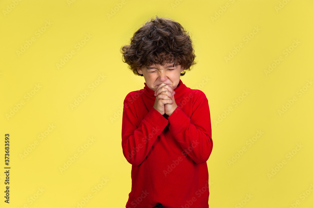 Praying. Portrait of pretty young curly boy in red wear on yellow studio background. Childhood, expression, education, fun concept. Preschooler with bright facial expression and sincere emotions. Praying. Portrait of pretty young curly boy in red wear on yellow studio background. Childhood, expression, education, fun concept. Preschooler with bright facial expression and sincere emotions.
